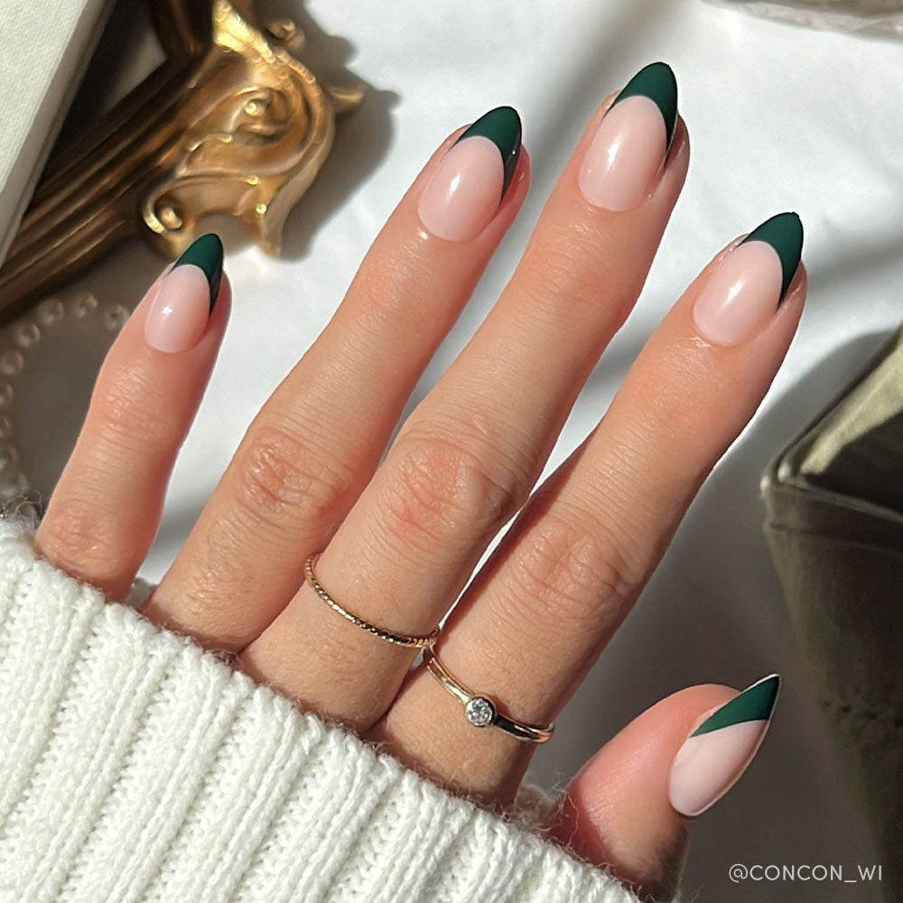 A hand with short almond nails showcases glamnetic’s Château Nail Set, featuring a modern dark green French tip on a nude base. Two delicate gold rings and a white knit sweater complete the chic look.