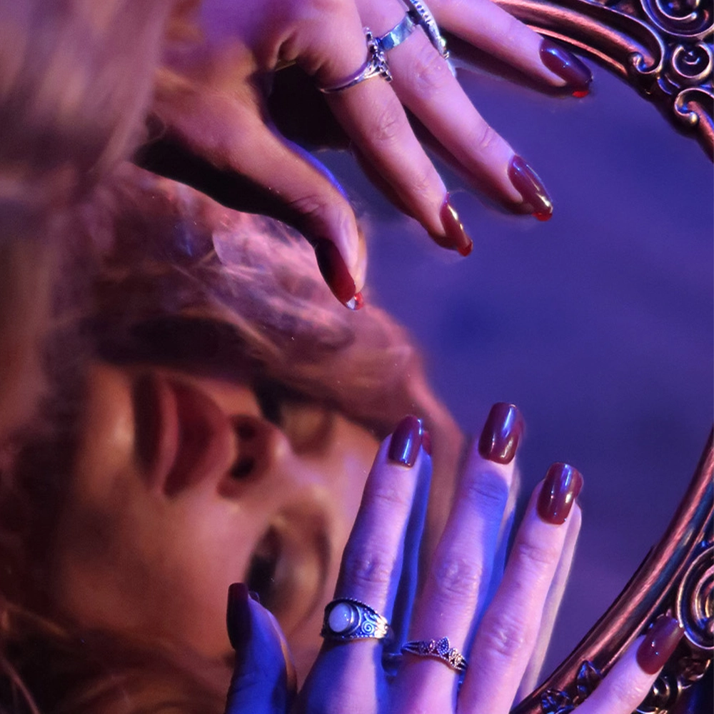 A womans face is reflected in an ornate mirror as her hands, featuring glamnetics Cherry Mocha gloss on medium square nails and adorned with rings, rest on the edge. Purple-blue lighting adds a dramatic, artistic touch. - Product image