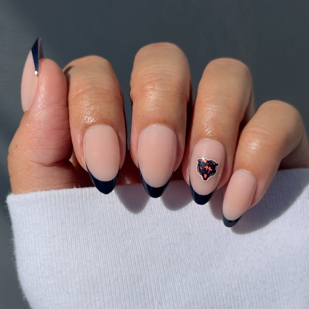 A hand with nude, almond-shaped nails featuring black French tips and a Chicago Bears logo on the ring finger displays the glamnetic Chicago Bears Press-On Nail Set, paired with a white long-sleeve top.