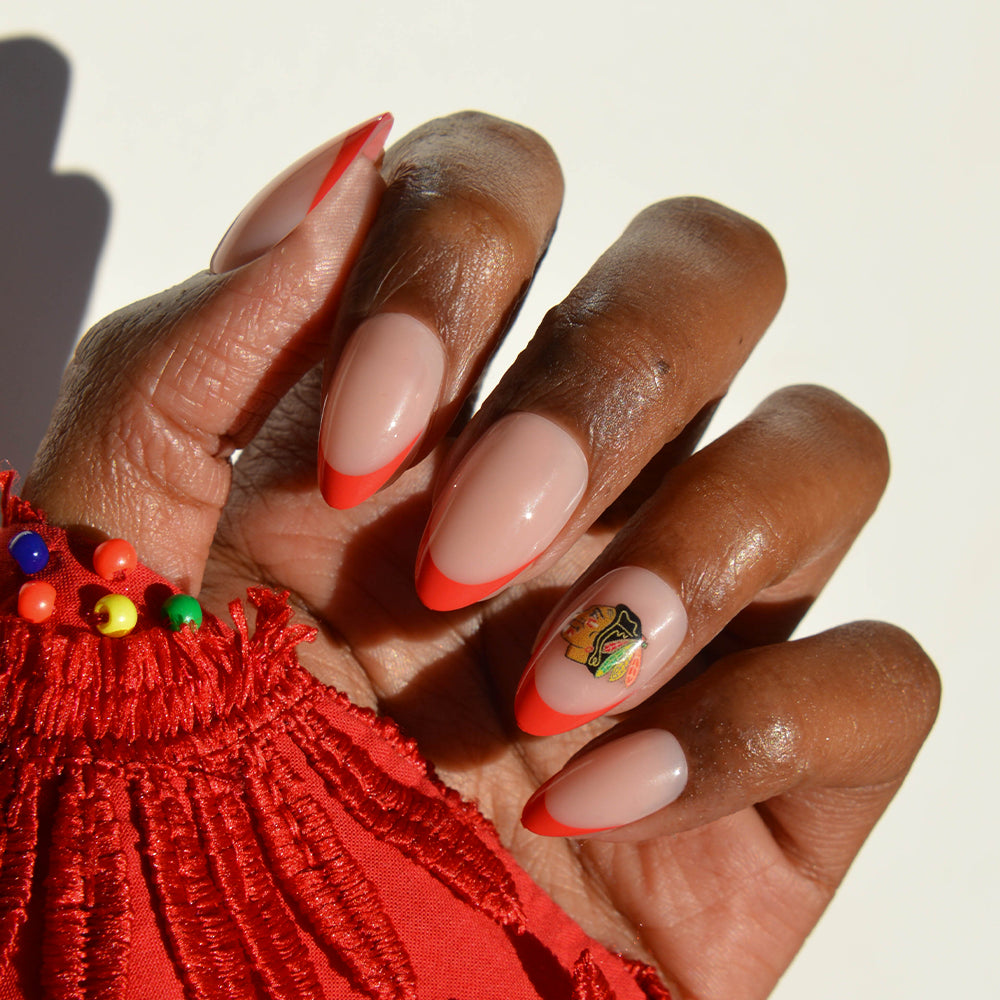 A hand with long, almond-shaped glamnetic NHL Press-On Nails showcases a red French tip design, accented by a small, colorful sticker on one nail. The person also wears a red fringed sleeve adorned with multicolored beads. - Product image