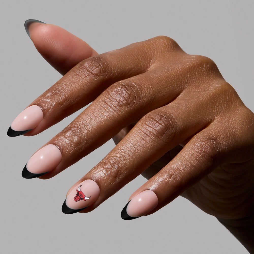 A hand with almond-shaped French tip press-on nails from the glamnetic Chicago Bulls Press-On Nail Set features a nude base, black tips, and a ring finger accent with a bold red Chicago Bulls logo.