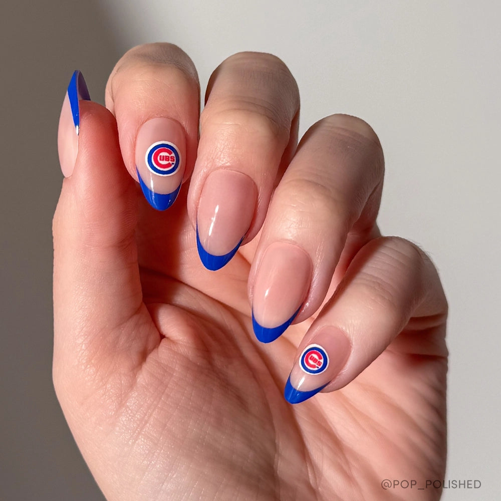 A hand wears glamnetics Chicago Cubs Press-On Nail Set, with almond-shaped nails and blue French tips. The ring and middle fingers feature a Chicago Cubs logo, shown on a plain, neutral background.