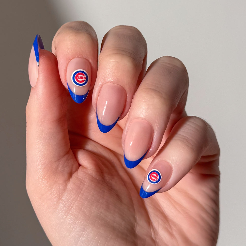 A hand with long nails, styled in a French tip with blue tips using glamnetic MLB Press-On Nails, features two accent nails adorned with small Chicago Cubs logo stickers near the cuticle. - Product image