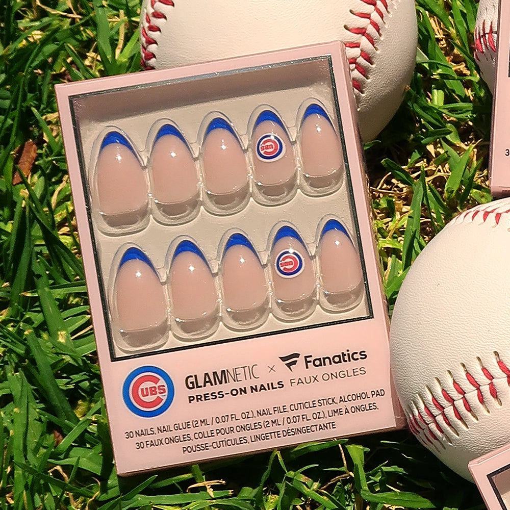 A box of glamnetics Chicago Cubs Press-On Nail Set is shown on grass with baseballs. The pink nails feature blue tips and the Cubs logo for a sporty, stylish look. - Product image