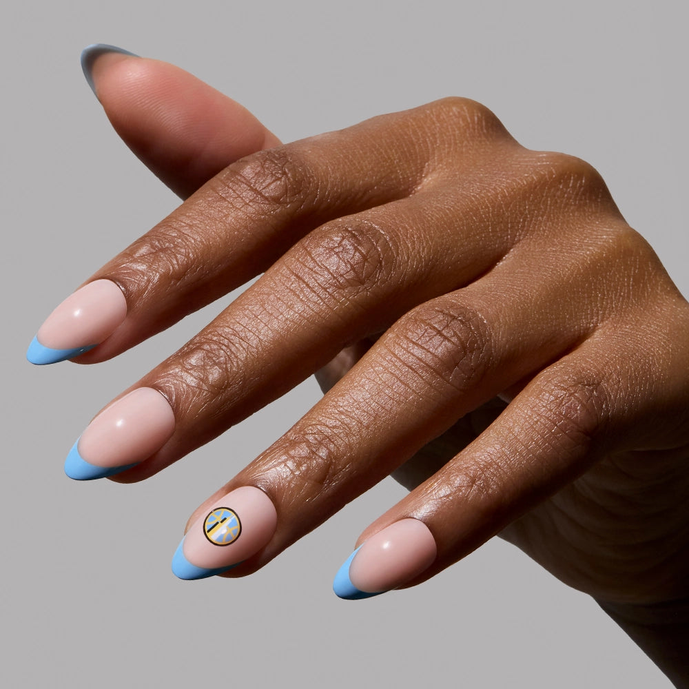 A hand with almond-shaped nails featuring a light pink base, pale blue French tips, and a small cross on the ring finger highlights a salon-quality mani using the glamnetic Chicago Sky Press-On Nail Set.