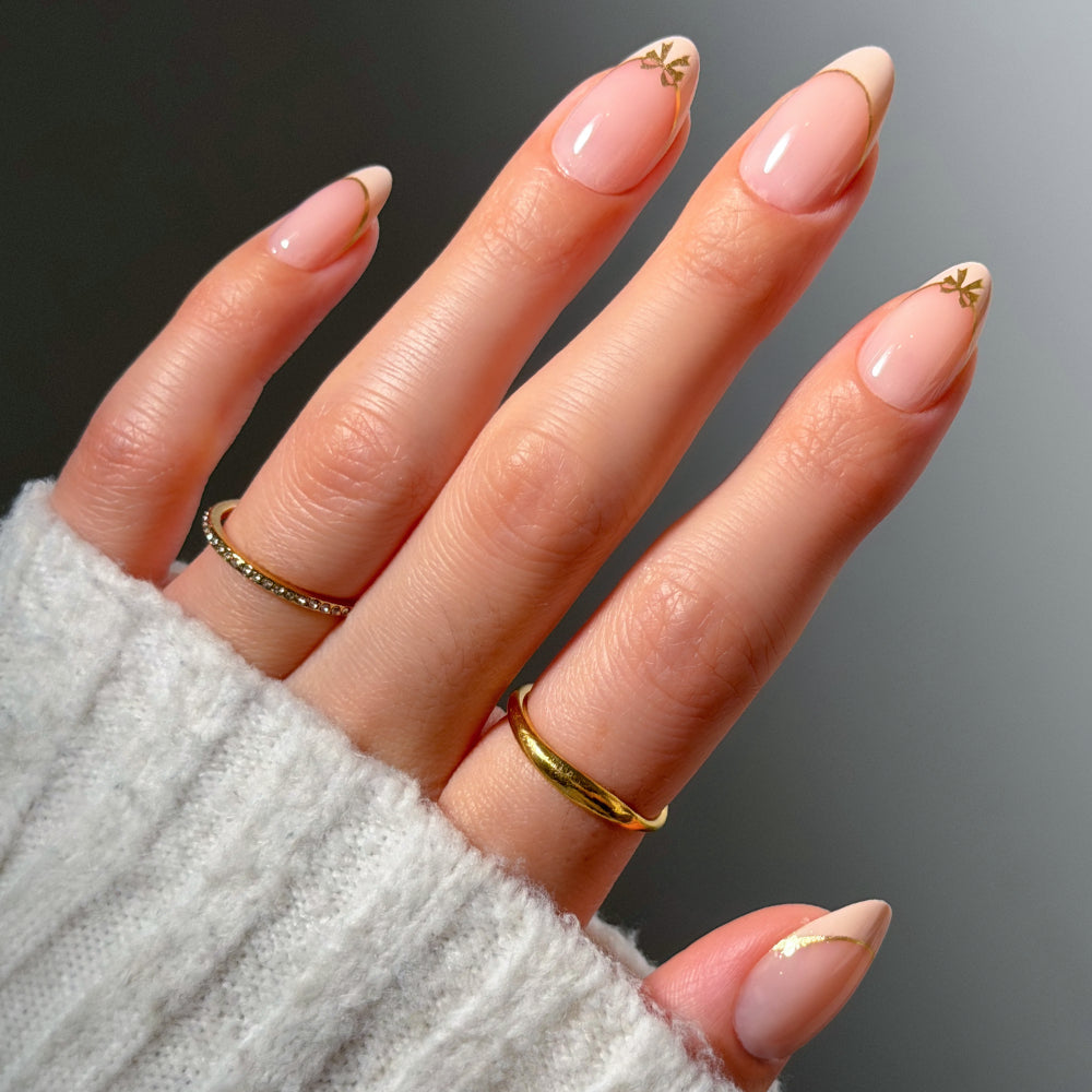 Glamnetic Cindy Lou press-on nails in a short almond shape with a sheer pink base, gold French tips, and small gold bow accents at the cuticles.