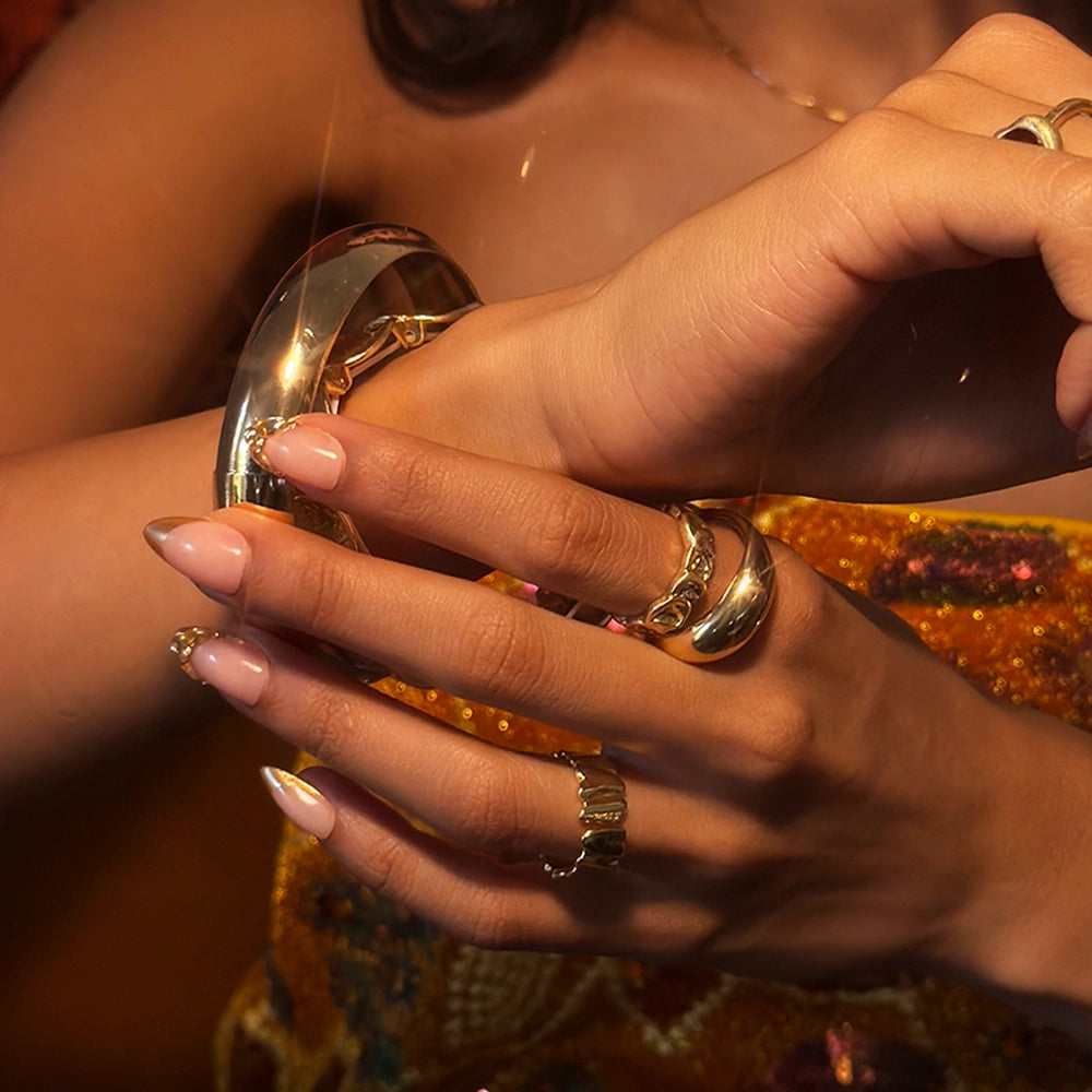 A close-up of a person’s hands with manicured almond nails, wearing multiple gold rings and holding the thick gold Citrine bracelet by glamnetic. The person is dressed in a sequined, gold-toned garment. - Product image