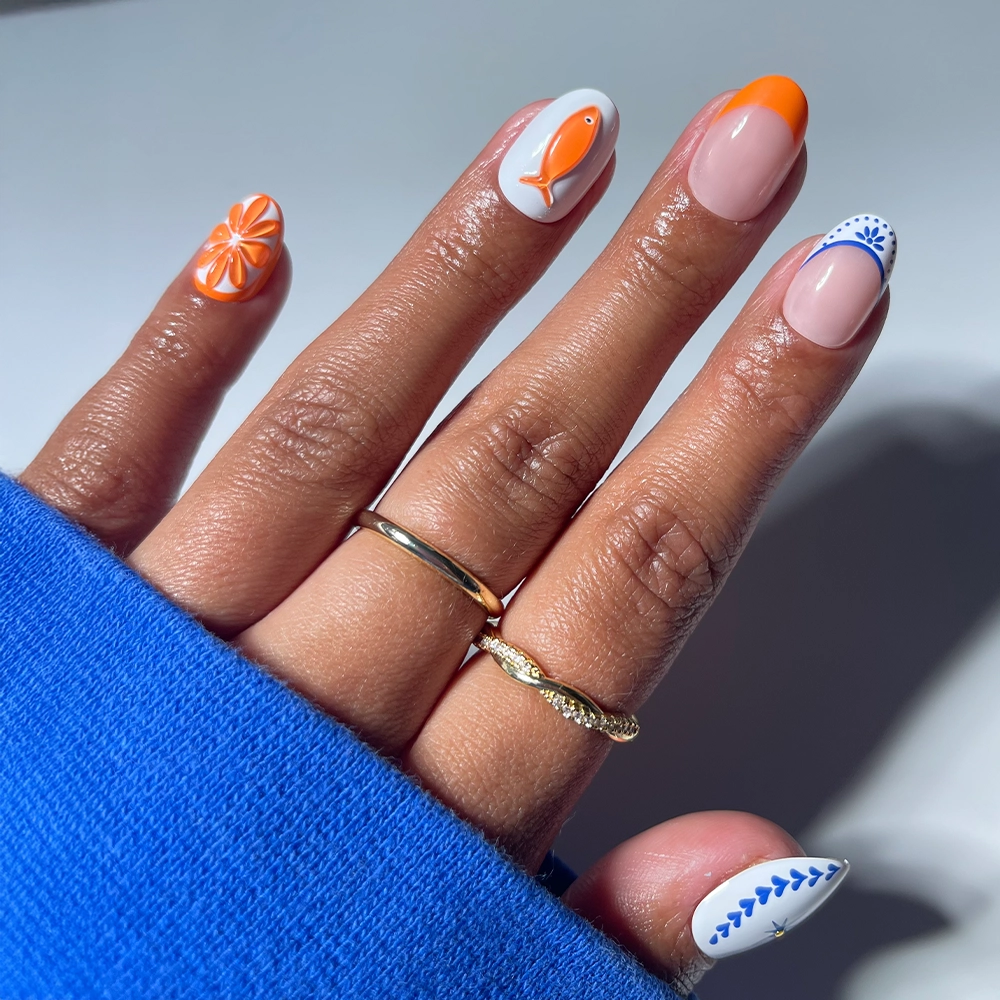 A hand with neatly manicured nails features glamnetic's Citrus Coast press-on set: short oval nails in creative designs—orange flower, fish, French tip, blue dotted tip, and leafy pattern—styled with three gold rings and a blue sweater.