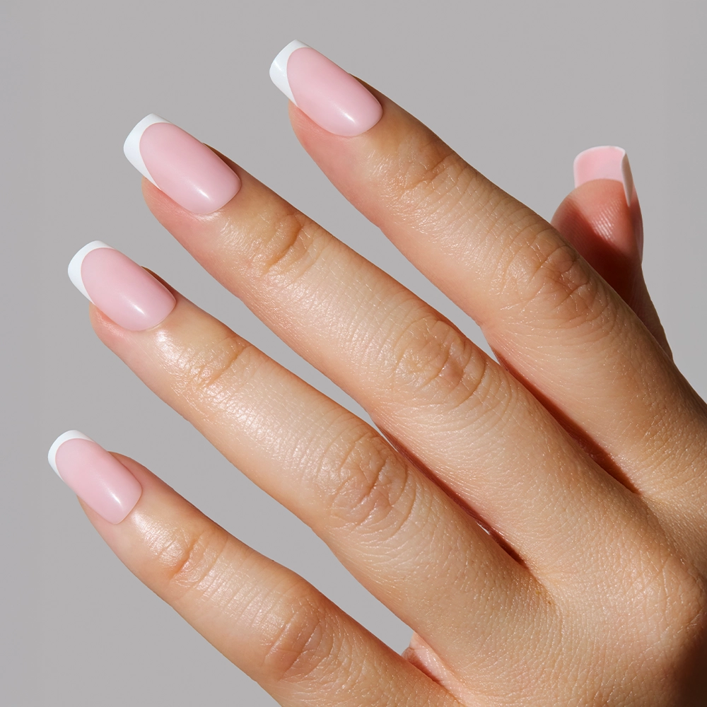 A close-up of a hand with glamnetic’s Classy French nails, featuring light pink nail beds and elegant white tips, presents a modern twist on the classic French manicure against a plain gray background. - Product image