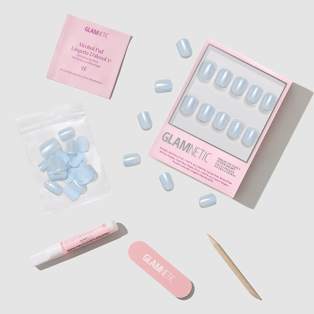 A flat lay of the glamnetic Clear Skies short press-on nails kit, with light blue salon-quality nails, nail file, cuticle stick, nail glue, alcohol pad, and extra nails neatly arranged on a light gray background. - Product image