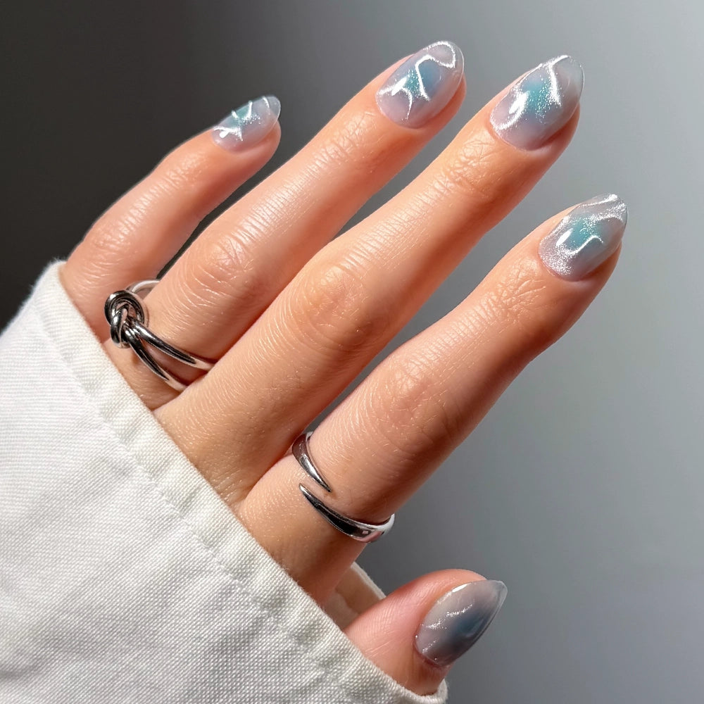 A close-up of a hand with almond-shaped nails painted in a iridescent silver and pale blue glitter manicure, wearing two silver rings, with a white sleeve visible. - Product image