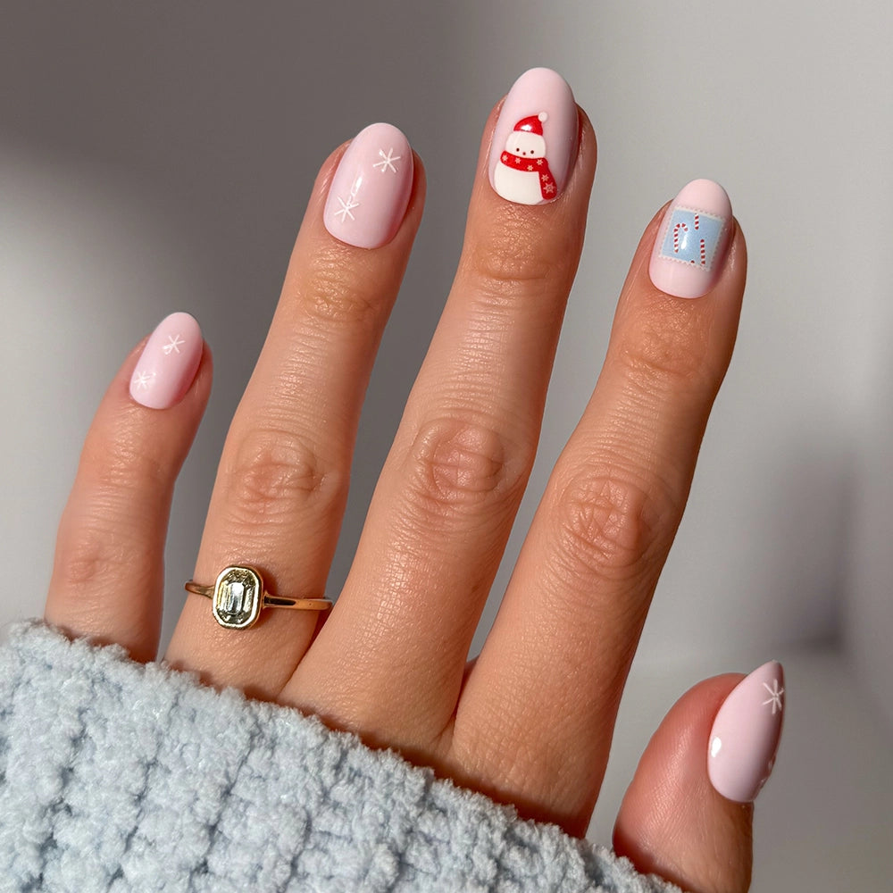 A hand with short oval nails in the shade Cocoa &amp; Kisses by glamnetic, featuring light pink polish with white stars and festive designs like a candy cane and a gift box. The person is wearing a gold ring and a cozy, light blue sweater.
