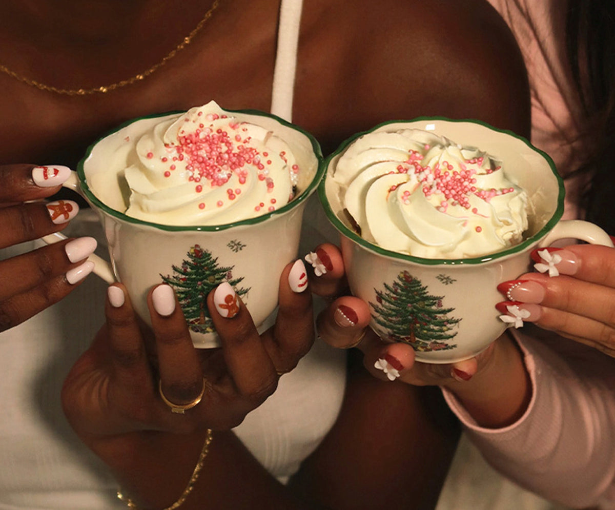 Two people holding mugs of hot chocolate with whipped cream and Christmas tree design.
