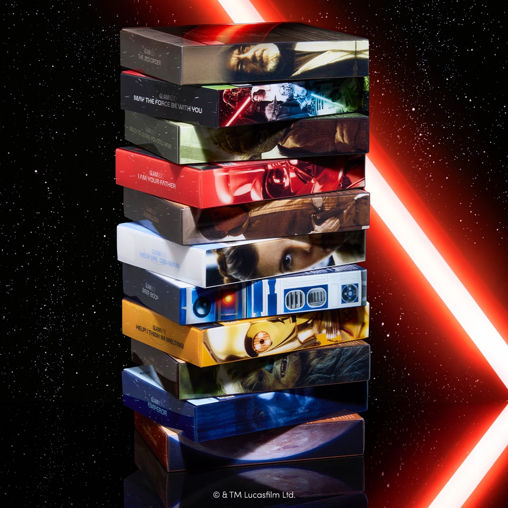 A stack of rectangular boxes featuring Star Wars characters and scenes, set against a starry backdrop with two red lightsaber beams crossing behind them—perfect for showcasing glamnetic’s cruelty-free and stylish STAR WARS press-on nails in the Emperor design. - Product image
