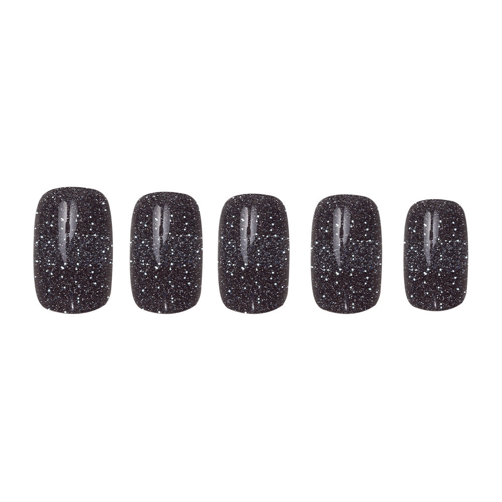 Five artificial nails from glamnetics Cosmic set display a semi-transparent black with silver glitter in a short squoval shape, arranged from largest to smallest against a white background. - Product image
