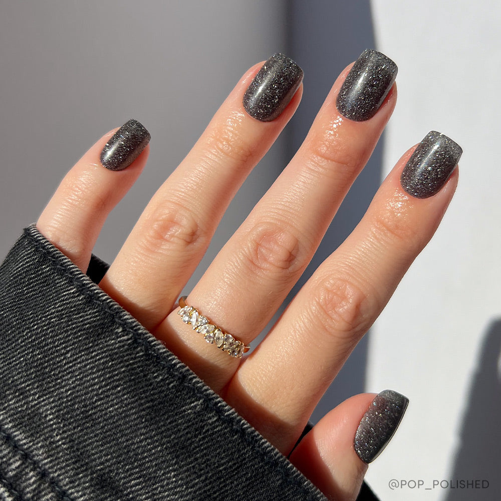A short squoval hand shows off glamnetic’s Cosmic Nail Set in a dark gray, semi-transparent black. Sunlight highlights the shimmer as the person, wearing a gold ring with clear stones and a black denim jacket, poses.