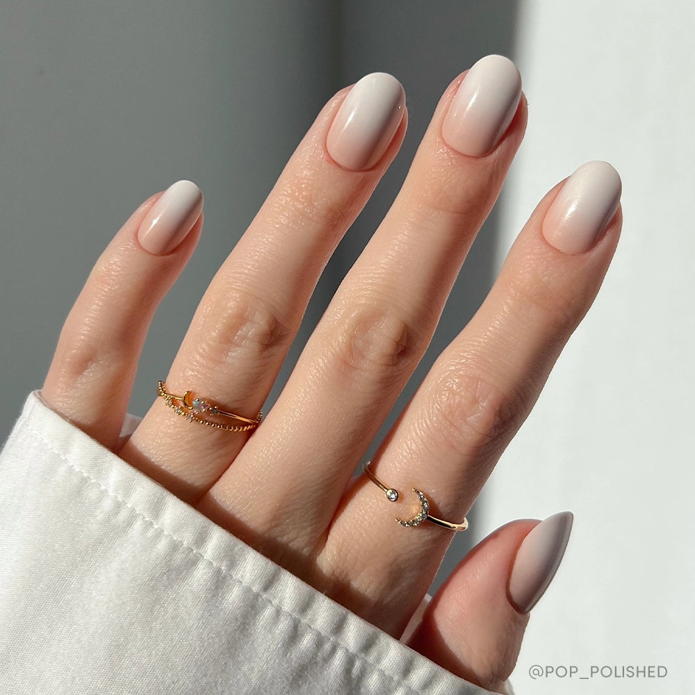 A hand with neatly manicured, short, almond-shaped vegan nails painted in Creamer by glamnetic—a light beige shade. The person wears two delicate gold rings with small gems. A white shirt cuff and a softly lit background complete the look. - Product image