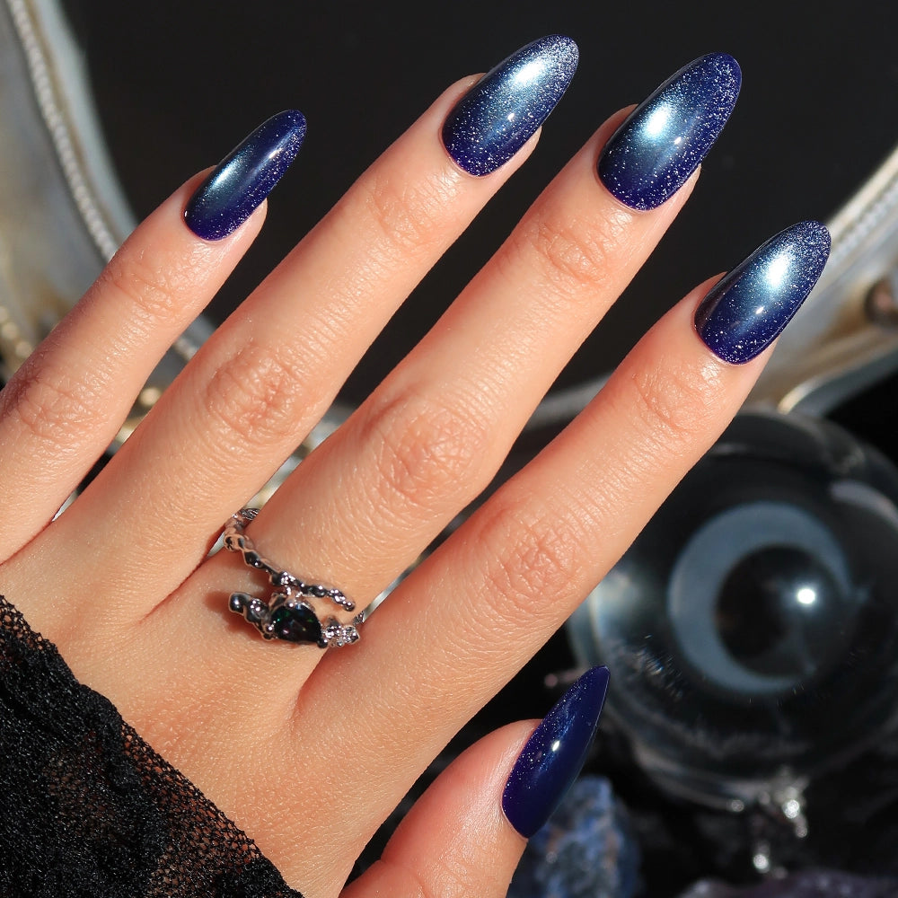 A hand with long, almond-shaped nails features glamnetic’s Crystal Cantation, showcasing a dark blue shimmer with a radiant gradient effect. The look is completed with a silver ring set with black stones and a black lace sleeve.