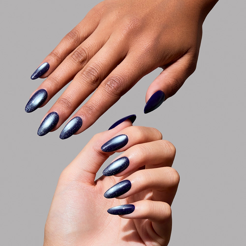 Two hands with almond-shaped, medium oval nails are displayed against a grey background. The press-on nails, Crystal Cantation by glamnetic, showcase a celestial deep blue ombre design with a shimmering, glittery finish that fades from dark navy at the tips to a lighter metallic blue center, creating an enchanting velvet effect. - Product image
