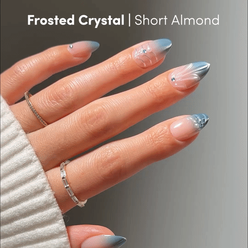 A short almond-shaped, salon-quality manicure—Frosted Crystals by glamnetic—shows off blue and silver frosted designs with tiny gem accents, paired with silver rings and a light knit sleeve.