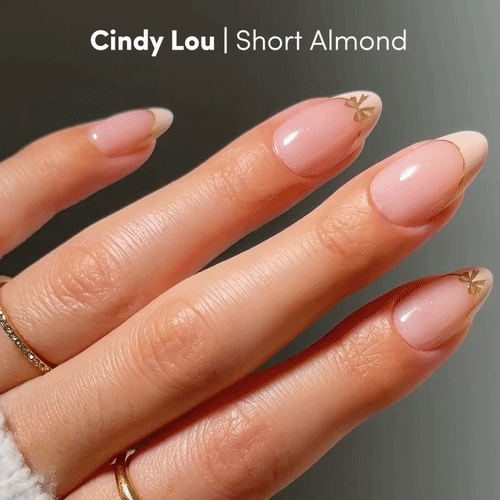 A hand with short almond-shaped nails features glamnetic Cindy Lou, a soft pink vegan polish with delicate gold accents near the tips. The fingers wear thin gold rings. Text reads: Cindy Lou | Short Almond vegan nails.