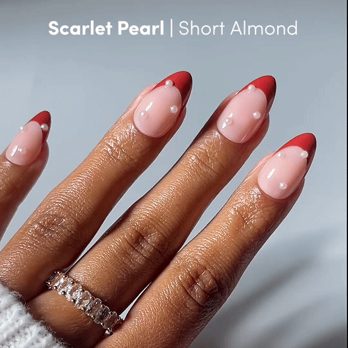 A hand with four nails painted in pink and red French tips with white dots shows off the Scarlet Pearl Press-On Nails by glamnetic, short squoval shape, paired with a silver ring with clear stones. Text: Scarlet Pearl | Short Squoval.