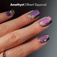 Close-up of a hand with short, squoval-shaped nails adorned in glamnetic's Amethyst press-on nails. The shimmering amethyst color with purple and pink hues is beautifully accented by gold trim and small gems, creating a luxurious velvet finish. Perfect for showcasing these elegant press-ons that make a stunning statement in any setting.