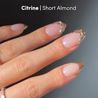 A hand displays short, almond-shaped press-on nails from glamnetic’s Citrine set: nude base color paired with glossy gold French tips. Text reads, Citrine | Short Almond.