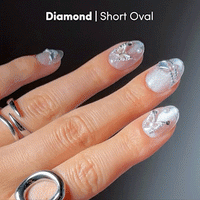 A hand with short oval nails, featuring glamnetic's Diamond press-ons in a shimmery silver shade, decorated with diamond-like accents and intricate 3D silver nail art. The look is complemented by silver rings. Text reads: Diamond | Short Oval.