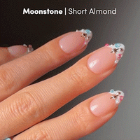 Close-up of a hand with short almond French tip nails featuring the "Moonstone" design by glamnetic, adorned with pastel beads, silver stars, and swirls on a pale nude base—ideal for achieving jewel-encrusted manicure elegance.