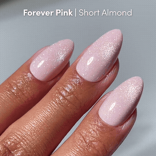 A hand with short almond glamnetic press-on nails in “Forever Pink,” a shimmery light pink, looks glossy and well-manicured against a soft gray background.