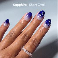 A hand adorned with a diamond ring showcases glamnetic's Sapphire Press-On Nails in the Short Oval shape, featuring a luxurious sapphire blue-to-light ombre gradient.