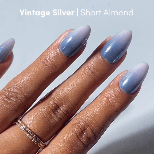 A hand with short, almond-shaped nails displays a gradient blue-to-silver ombre manicure using glamnetic’s Vintage Silver Press-On Nails. The text reads: “Vintage Silver | Short Almond.” The hand is adorned with a delicate, gold double-band ring. - Product image
