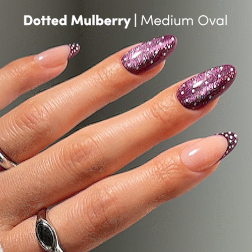 A hand with medium oval nails from glamnetic’s “Dotted Mulberry” set, featuring playful mulberry-colored glitter French tips and dotted accents that create a chic polka-dot design. The text reads: Dotted Mulberry | Medium Oval. Silver rings adorn the hand, highlighting this press-on nail set’s stylish purple details. - Product image