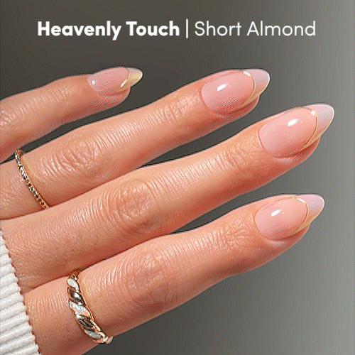 A hand with short almond nails from glamnetic’s “Heavenly Touch” set, showcasing a pink and nude gradient with gold tips. The person wears a gold ring and a white ribbed sleeve. Text reads: Heavenly Touch | Short Almond — perfect for anyone seeking elegant press-on nails by glamnetic. - Product image