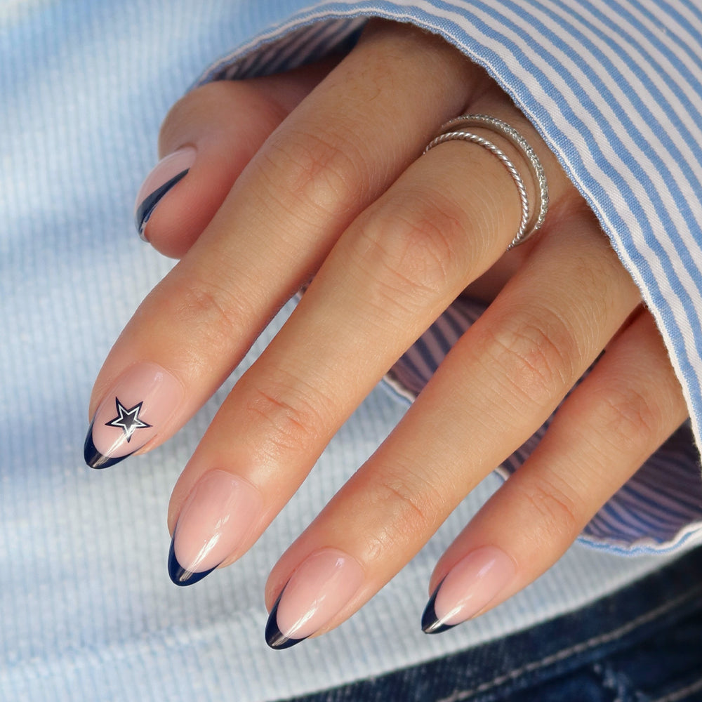 A hand featuring almond-shaped NFL Press-On Nails by glamnetic, styled with nude polish, thin navy blue tips, and a small star design on the ring fingernail. The person is wearing a silver ring and a blue striped sleeve. - Product image