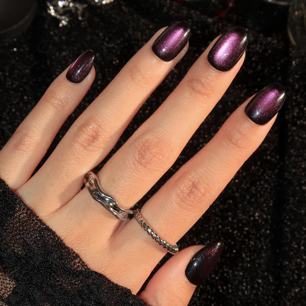 A hand showcases glamnetic’s “Dark Romance” press-on nails, featuring a long, almond shape and a glossy, deep purple velvet finish. The look is complemented by two silver rings and a black mesh sleeve against a glittery black fabric background.