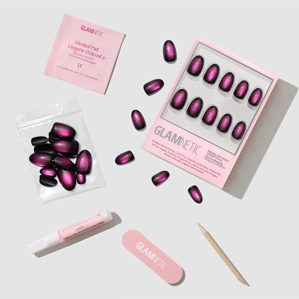 Introducing the Glamnetic Dark Romance press-on nails kit, showcasing short, oval-shaped nails in a gorgeous purple-black ombré with a luxurious velvet finish. The set includes an alcohol pad, nail glue, pink nail file, wooden cuticle stick, and a bag of extra nails—all elegantly arranged on a light surface. - Product image