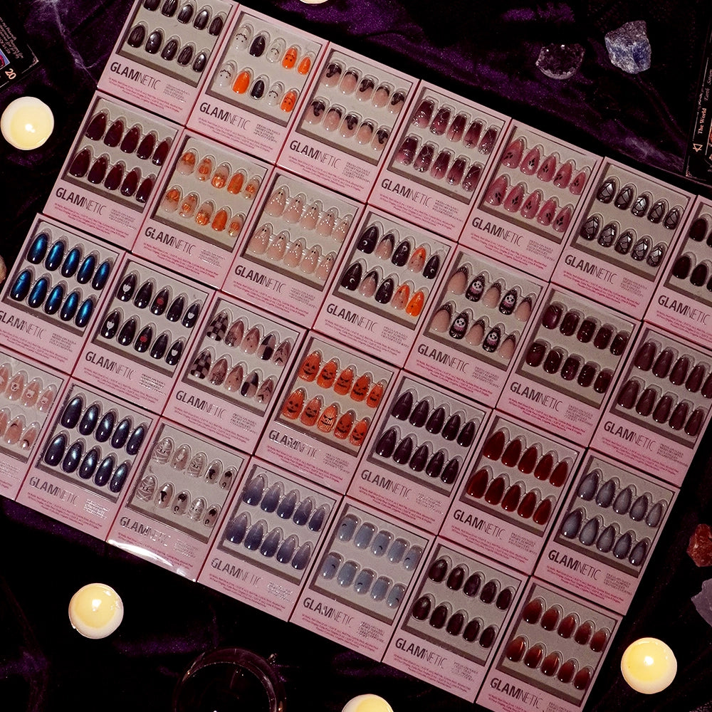 A display of glamnetic’s Dark Romance press-on nails in colorful packaging arranged neatly on a dark surface, surrounded by lit candles and decorative stones. Featuring both vibrant patterns and elegant short oval nails with a velvet finish, this arrangement showcases stylish options for every occasion. - Product image
