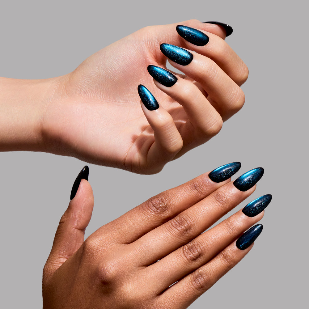 Two hands with different skin tones display long, almond-shaped Glamnetic press-on nails in the shade “Dark Waters,” painted with a shimmering, metallic teal cat-eye polish against a plain gray background, highlighting the convenience and beauty of these long-lasting nails. - Product image