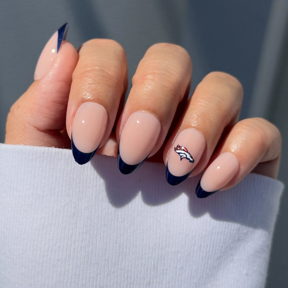 A hand sports glamnetic’s Denver Broncos Press-On Nail Set: almond-shaped nude nails with dark blue French tips and a signature Broncos logo accent. The look is paired with a white long-sleeve shirt—perfect for fans.