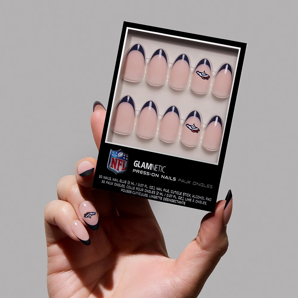 A hand holds a box of glamnetic Denver Broncos Press-On Nails, featuring nude bases, dark tips, and a small team logo. The matching almond manicure is perfect for fans who love showing off their Denver Broncos nail art.