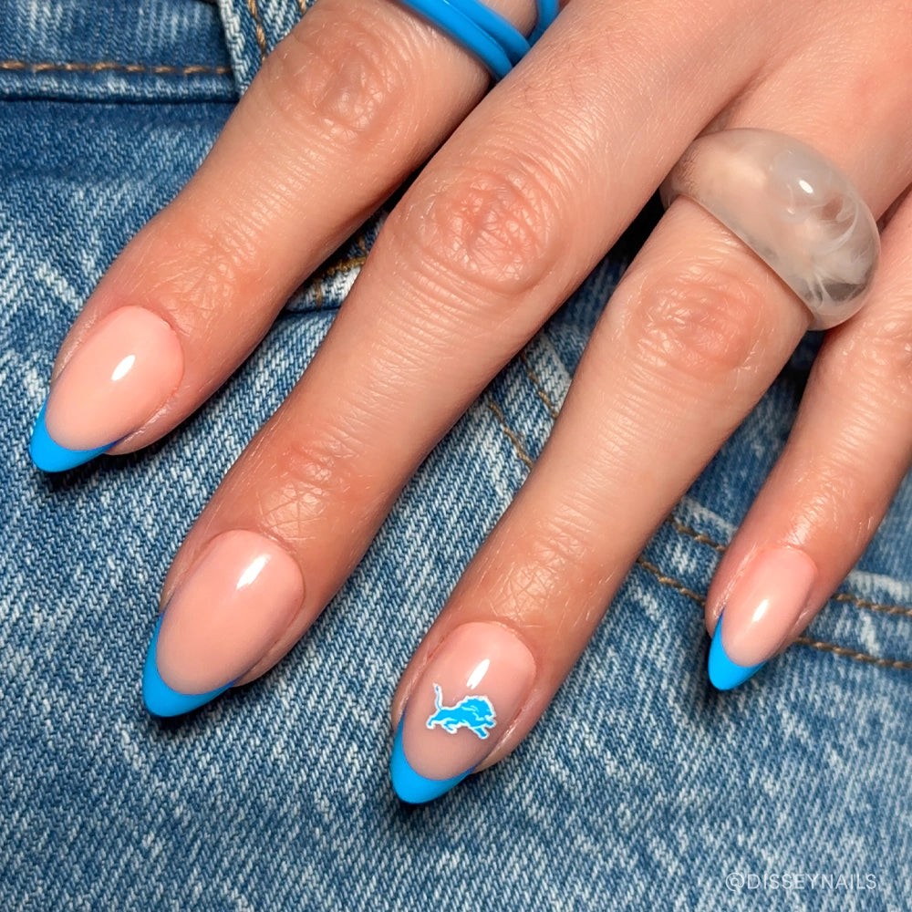 The glamnetic Detroit Lions Press-On Nail Set features almond-shaped nude nails with bright blue French tips and a signature logo accent—ideal for football fans. Displayed on blue denim, the look is finished with two translucent rings.