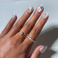 A hand with glamnetic's Diamond 3D butterfly nail art on short oval nails, paired with two gold rings—one featuring a gemstone and the other set with small diamonds—against a light background.