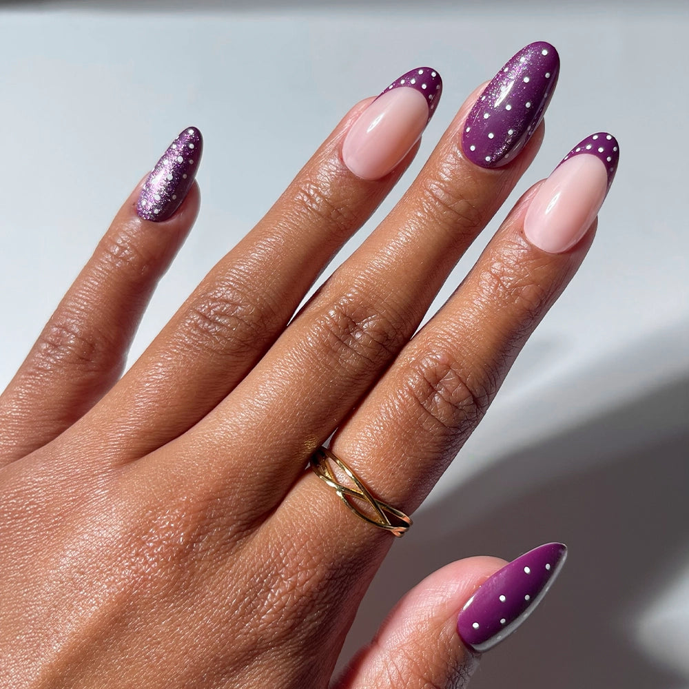 A hand showcases glamnetic’s Dotted Mulberry nails, featuring an almond shape with a purple gradient, playful white polka-dots, and chic nude tips. The look is completed with a gold twisted ring adorning the middle finger. - Product image
