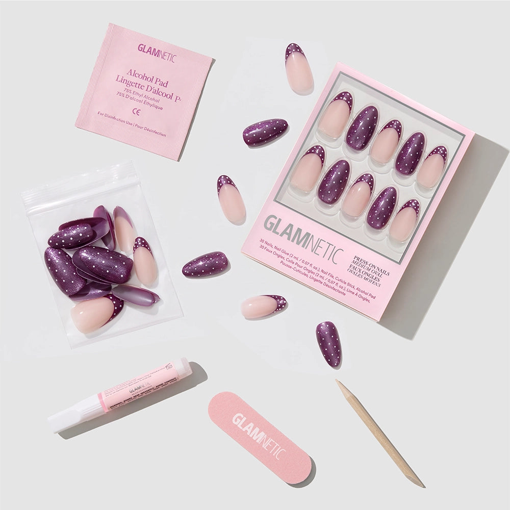 The Glamnetic Dotted Mulberry press-on nail set features purple and pink nails with playful polka-dot designs, and comes with glue, a wooden stick, nail file, alcohol pad, and extra nails arranged on a light gray surface. - Product image