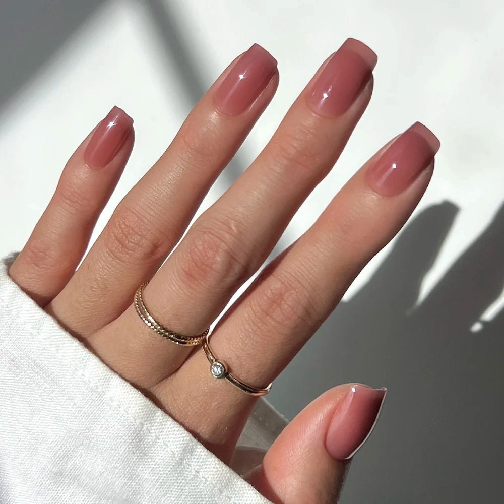 A hand with long, medium square nails in a semi-transparent, glossy nude-pink shade—wearing two gold rings—is shown against a soft white background, featuring the Glamnetic Dusty Pink press-on nails.