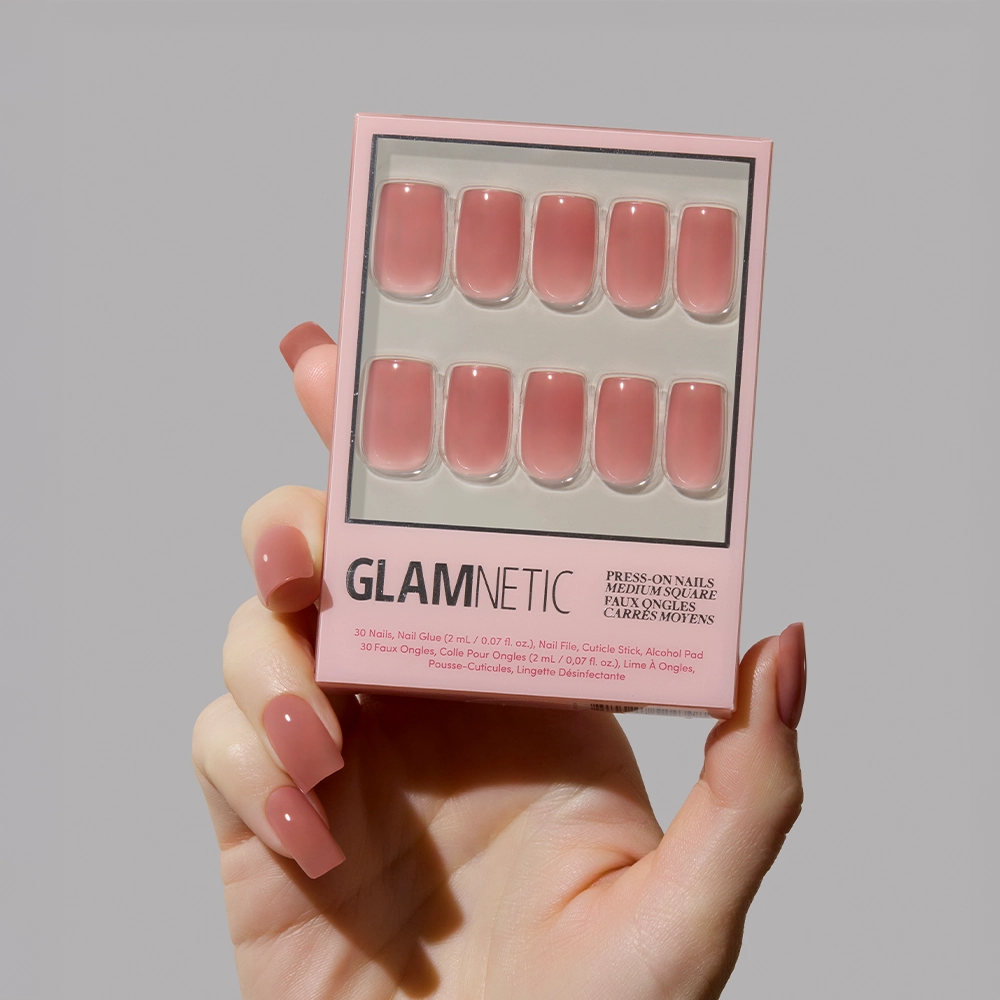 A hand with long, square-shaped nails in the glamnetic Dusty Pink shade holds the Dusty Pink press-on nails box against a plain light gray background—moody yet refined.