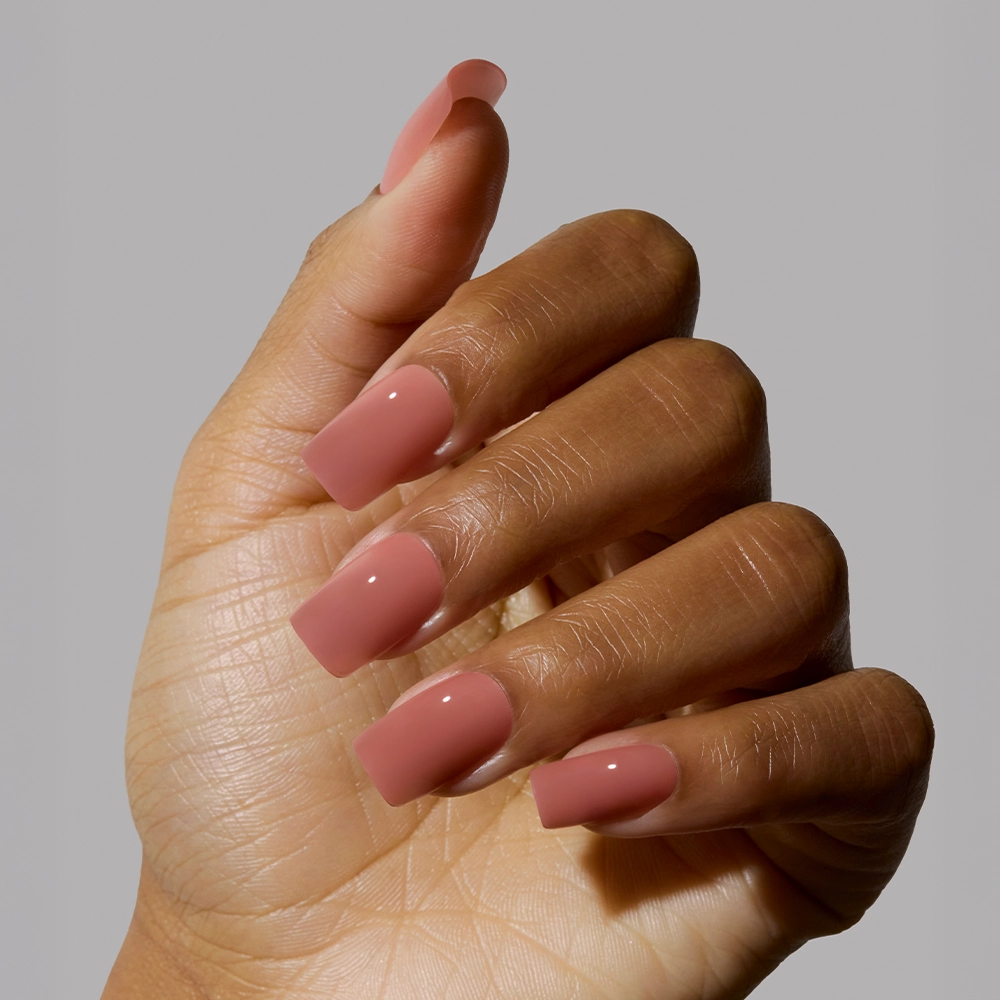 A hand with medium-dark skin tone shows long, medium square nails painted in glamnetics glossy Dusty Pink shade against a plain gray background. - Product image