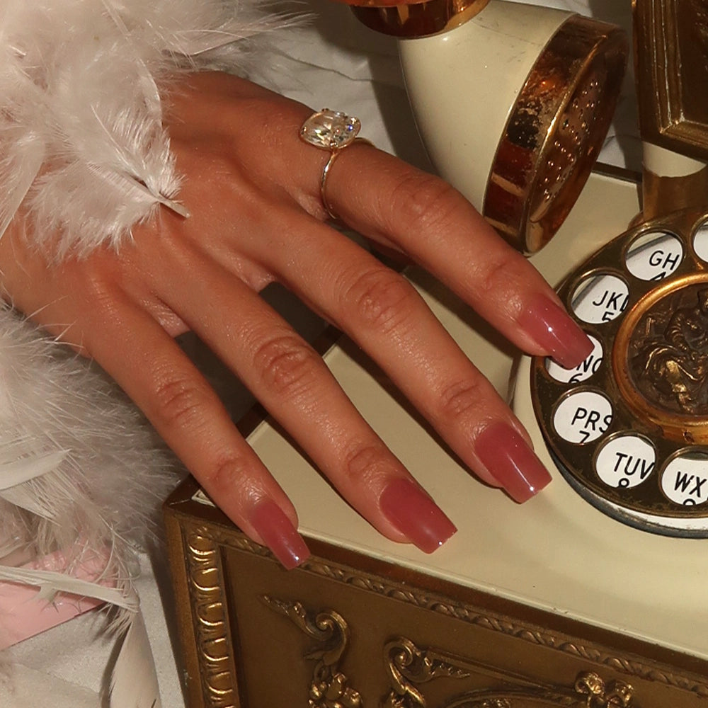 A hand with long, medium square glamnetic Dusty Pink nails and a glossy finish, wearing a large gemstone ring, rests on a vintage rotary phone next to a white feathered garment. - Product image